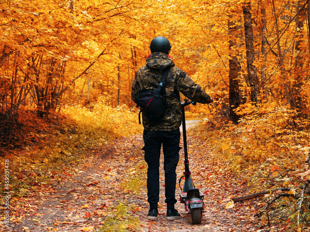 Naklejka premium A young man in the autumn forest on an electric scooter. Ride colorful autumn on forest roads.
