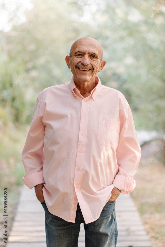 Portrait of a mature man looking at the camera and smiling while posing with his hands in his pockets outdoors in nature.