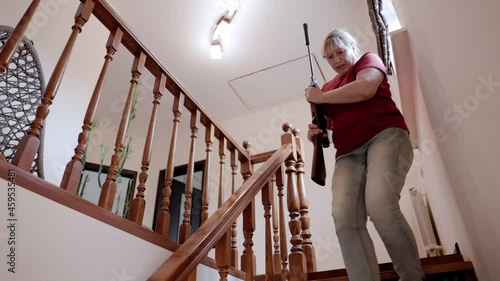 Caucasian woman in a red T-shirt descends the stairs with a rifle in her hands. Self defense concept. Middle aged woman holding a riffle.