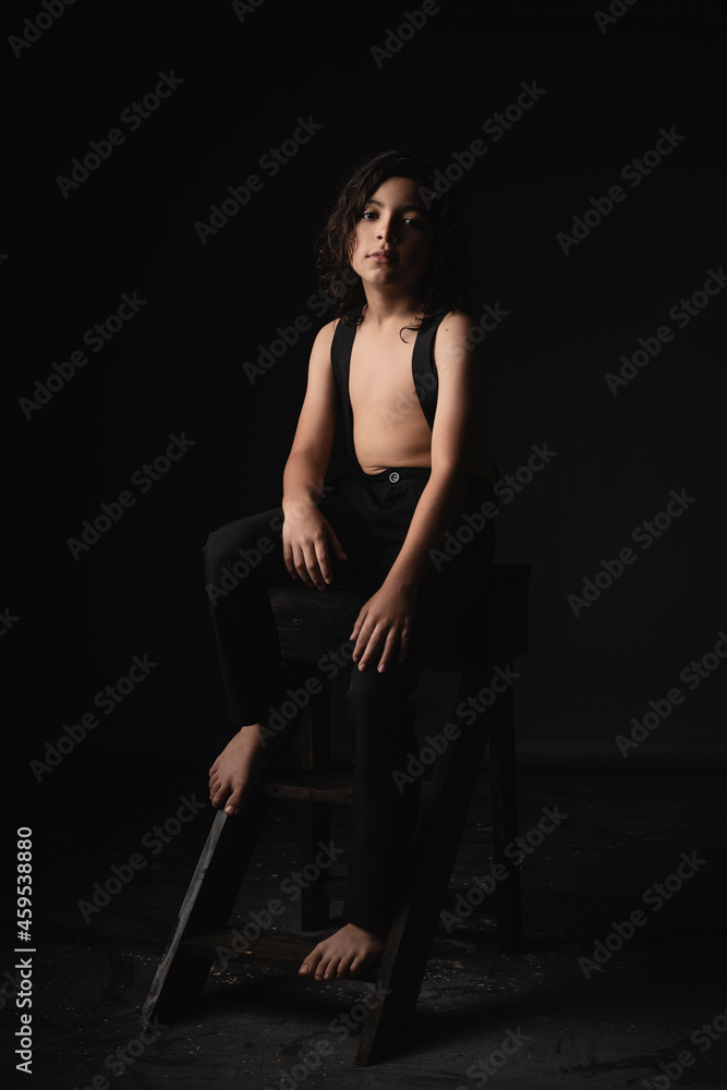 Fototapeta premium Niño de cabello largo sentado posando en estudio con fondo negro