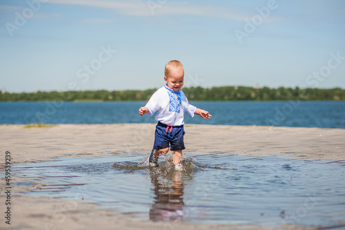 Toddler boy running through puddles close-up and copy space. Baby in embroidered shirt plays with water, Ukraine, Dnipro... © Nelly