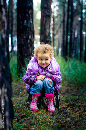 The little girl sits on the stump in the woods. Curly girl in rubber boots. The child slyly smiling.