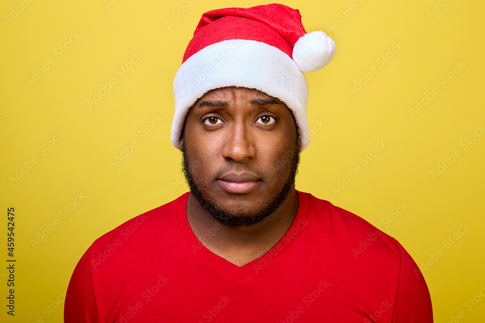 Portrait of an attractive black guy in a Christmas hat and a red T-shirt confidently looking directly into the camera isolated on a bright yellow background Portrait of an attractive black guy in a Christmas hat and a red T-shirt confidently looking directly into the camera isolated on a bright yellow background