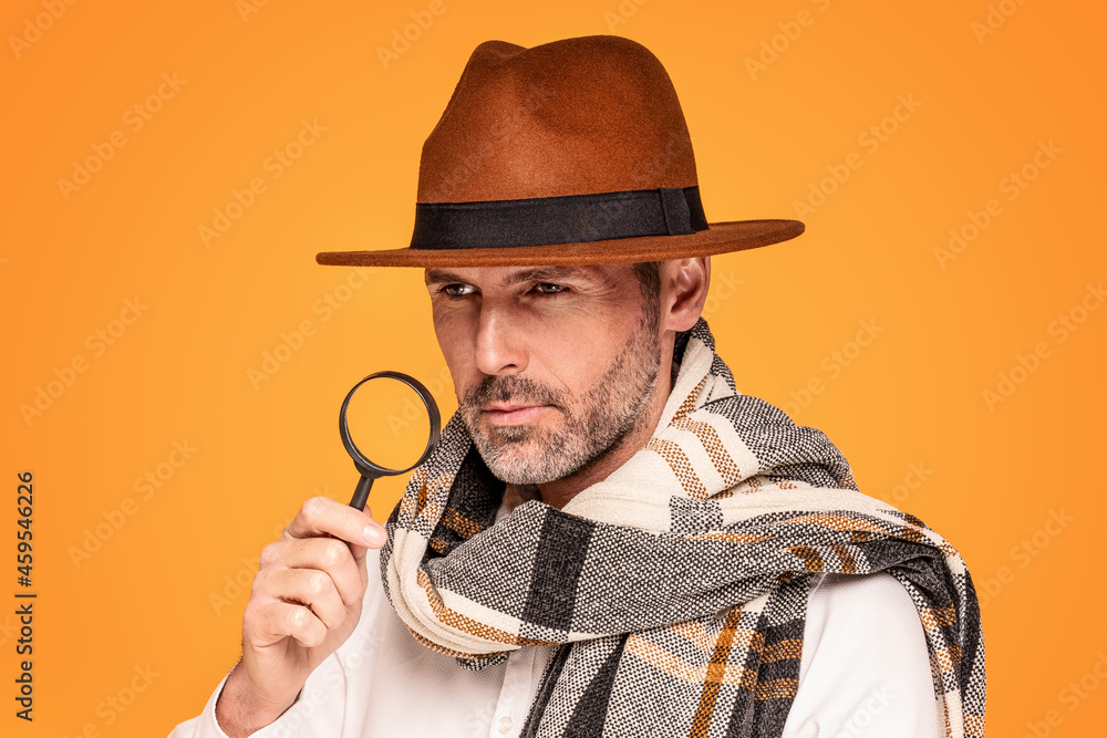 Male detective with magnifying glass, wearing fashionable scarf and brown hat, posing on yellow studio background. Male detective with magnifying glass, wearing fashionable scarf and brown hat, posing on yellow studio background.
