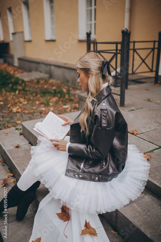 Blonde ballerina in white dress and black jacket reading a book while sitting on the steps of the street in autumn