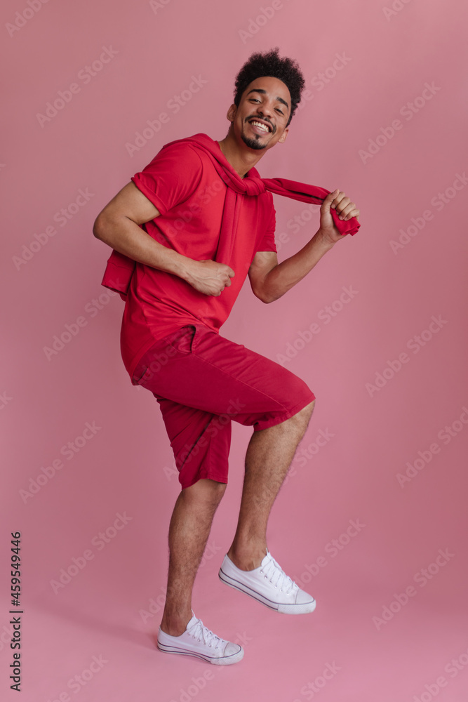 Young African American man in full-length studio shot is standing on one leg and holding sleeves of sweater. Cute dark-haired volleyball player with smile, dressed casual clothes. Happy days concept Young African American man in full-length studio shot is standing on one leg and holding sleeves of sweater. Cute dark-haired volleyball player with smile, dressed casual clothes. Happy days concept