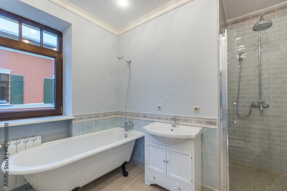 The bathroom is in blue tones with a window, a bathtub on curved legs, a sink and a shower cabin. Interior of a cottage or townhouse The bathroom is in blue tones with a window, a bathtub on curved legs, a sink and a shower cabin. Interior of a cottage or townhouse