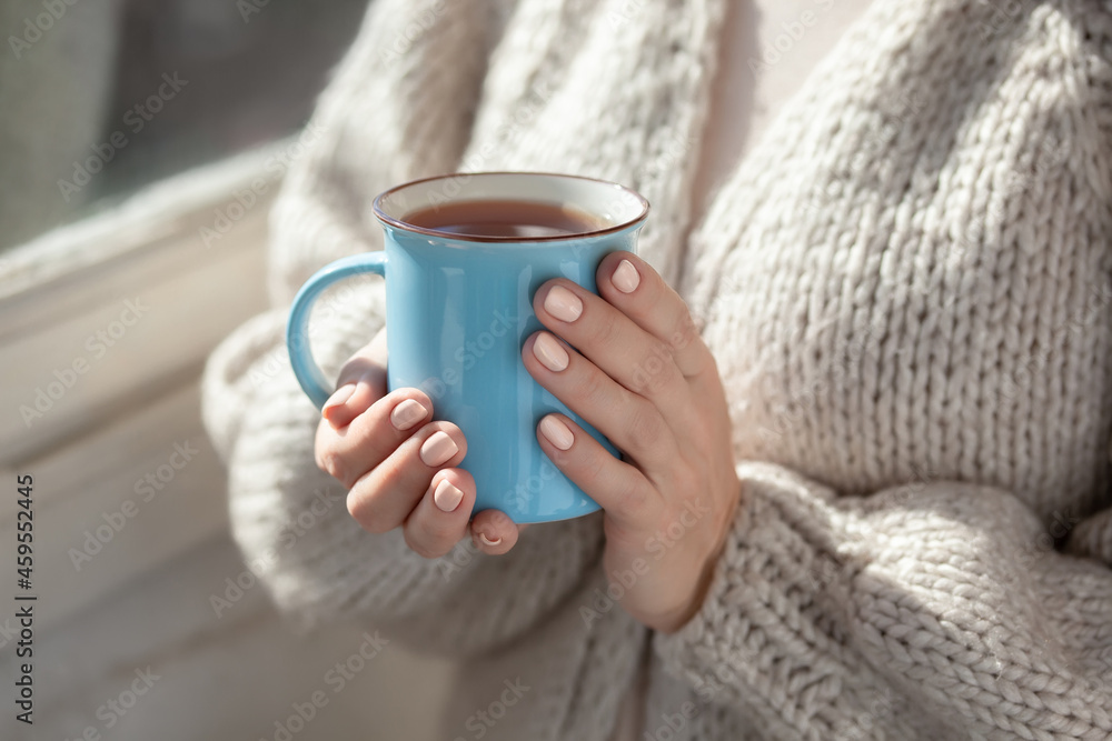 A woman in a warm cardigan holds a blue mug in her hands. Close-up of female hands holding cup of coffee or tea.