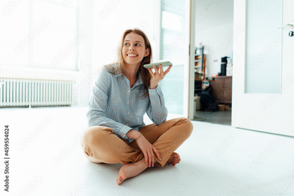 Smiling young woman talking on smart phone through speaker while sitting cross-legged at home Smiling young woman talking on smart phone through speaker while sitting cross-legged at home