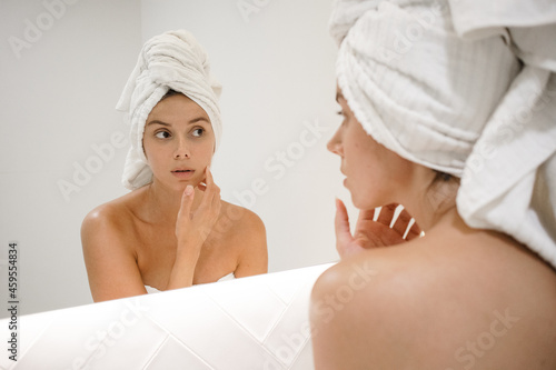 young white woman in the mirror towel on head looking at skin looking for spots signs of age and lines worried and surprised in the bathroom after shower self care skin care self love acne spots aging