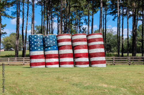 American flag painted on hay bales in a farm field against a backdrop of pine trees and blue sky. Patriotic theme.