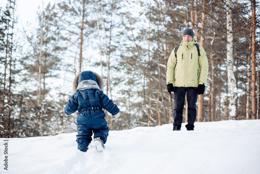 Little son happily going towards his dad. Father and baby playing in winter forest outdoors. Man with backpack and toddler boy in blue overalls walking in snowy nature park.