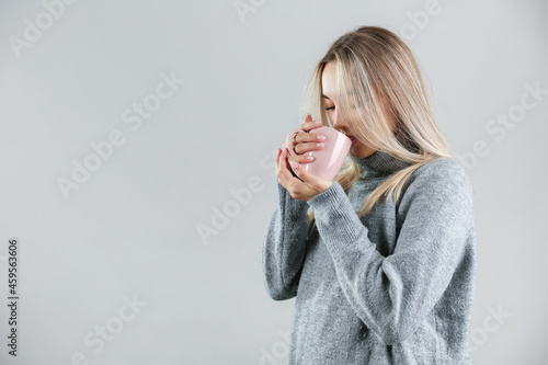 woman wearing warm sweater drinks a hot tea on grey background, studio shot. the concept of warming up with a hot drink. 