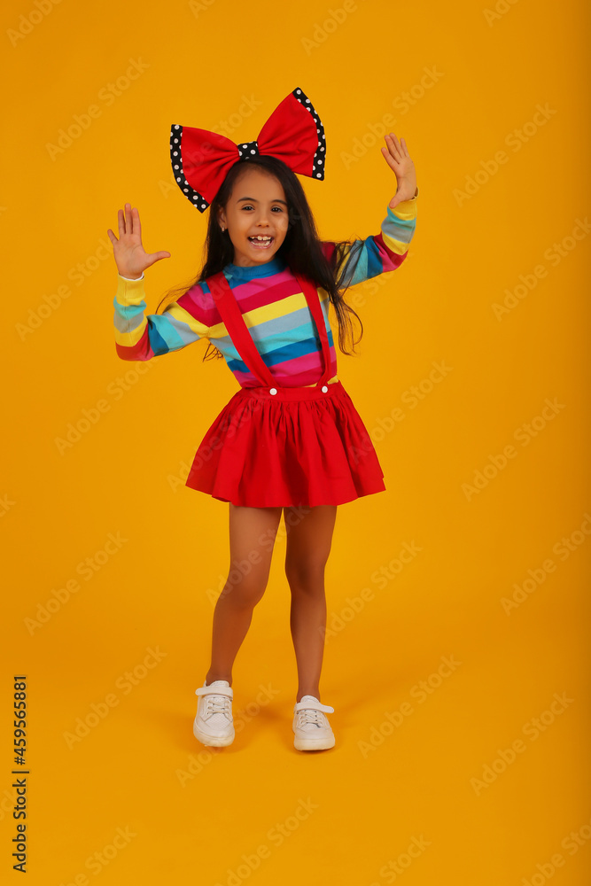 a dark-skinned happy girl in a red skirt in a colored jacket with a red polka dot bow on her head on a yellow background a dark-skinned happy girl in a red skirt in a colored jacket with a red polka dot bow on her head on a yellow background