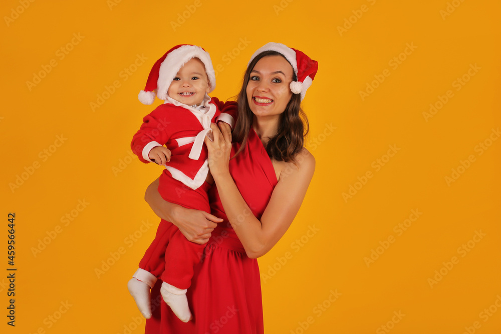 Fototapeta premium a beautiful young girl in a red dress holds her son in a Santa Claus costume and a Santa Claus hat for the new year