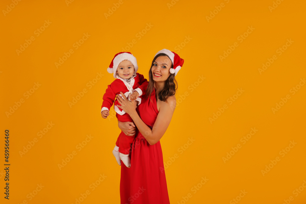 Obraz premium a beautiful young girl in a red dress holds her son in a Santa Claus costume and a Santa Claus hat for the new year