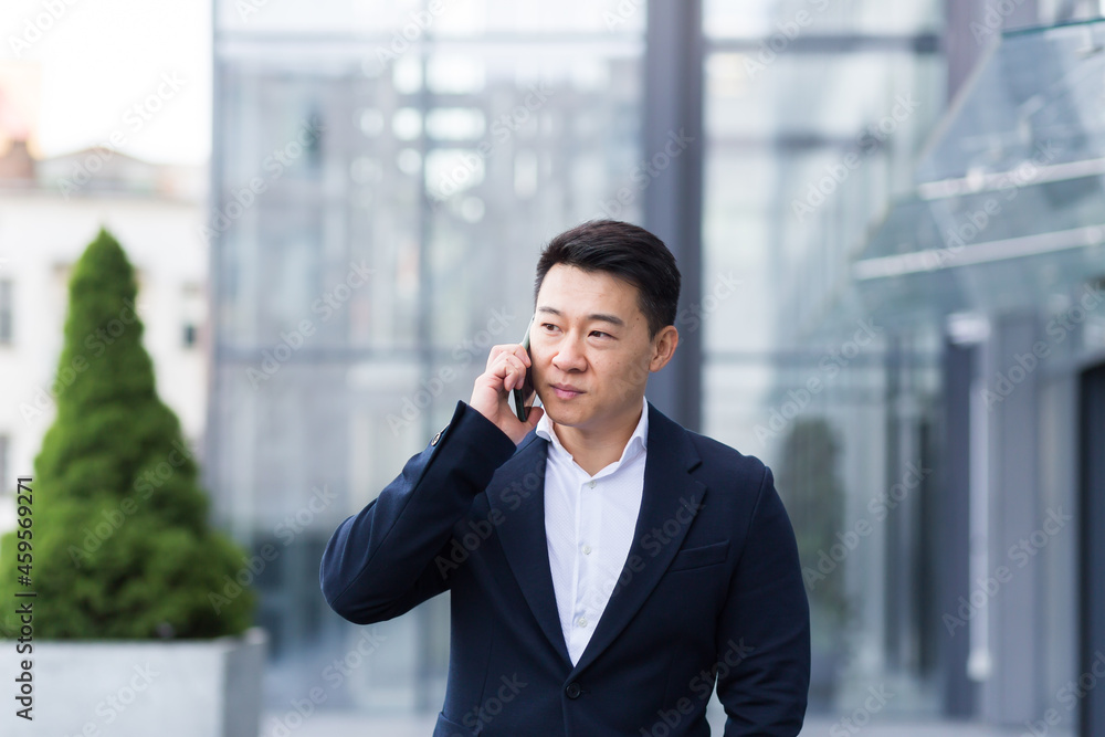 Successful Asian businessman businessman talking on a mobile phone while walking near the office of a large modern, serious and pensive employee Successful Asian businessman businessman talking on a mobile phone while walking near the office of a large modern, serious and pensive employee