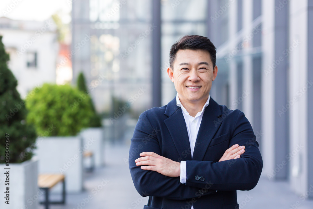 Successful Asian boss near the office looks at the camera with crossed arms smiles and rejoices in success Successful Asian boss near the office looks at the camera with crossed arms smiles and rejoices in success