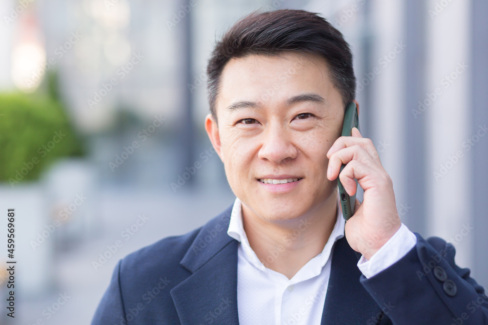 Successful asian businessman businessman talking on cell phone while walking near big modern office, cheerful smiling worker and looking at camera Successful asian businessman businessman talking on cell phone while walking near big modern office, cheerful smiling worker and looking at camera
