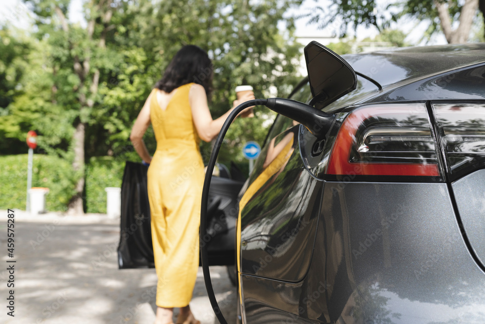 Car charging at electric station with woman holding coffee cup in background Car charging at electric station with woman holding coffee cup in background
