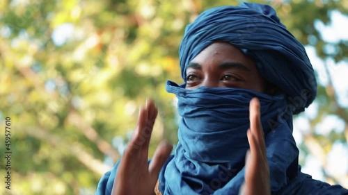 Man in traditional blue Tuareg dresses, sitting in a park listening to music and clapping with his hands. Blurred background of trees.
