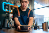 Asian man barista making hot coffee latte in coffee cup to customer on bar counter at cafe. Male coffee shop waiter serving hot coffee with milk to client. Small business restaurant owner concept.