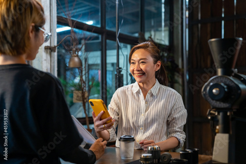 Asian woman customer using smartphone scanning bar code make contactless payment to barsita at cashier counter in coffee shop. Small business with modern mobile banking electronic payment concept