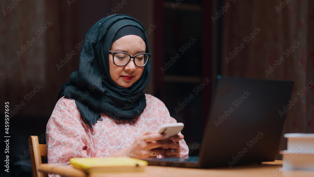 An office worker for an Asian Muslim woman sitting in front of a laptop computer at her desk and talking on a mobile phone and working at the office. An office worker for an Asian Muslim woman sitting in front of a laptop computer at her desk and talking on a mobile phone and working at the office.