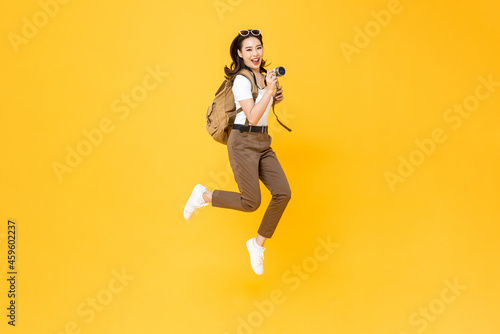 Young pretty Asian woman tourist backpacker smiling and jumping with camera in hand isolated on yellow studio background