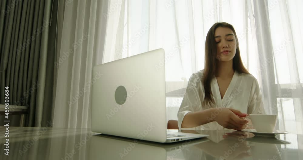 Pretty asian girl in white robe drinking coffee while using laptop in early morning, surfing internet on computer. Gen z girl remotely working from vacation in hotel 4k footage Pretty asian girl in white robe drinking coffee while using laptop in early morning, surfing internet on computer. Gen z girl remotely working from vacation in hotel 4k footage