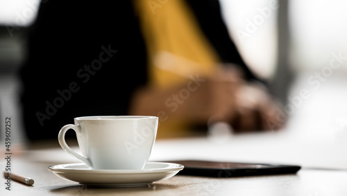 Close up shot of white hot coffee cup and small dish on meeting table near pencil while unidentified unrecognizable businessperson take short break after discussing and planning in blurred background