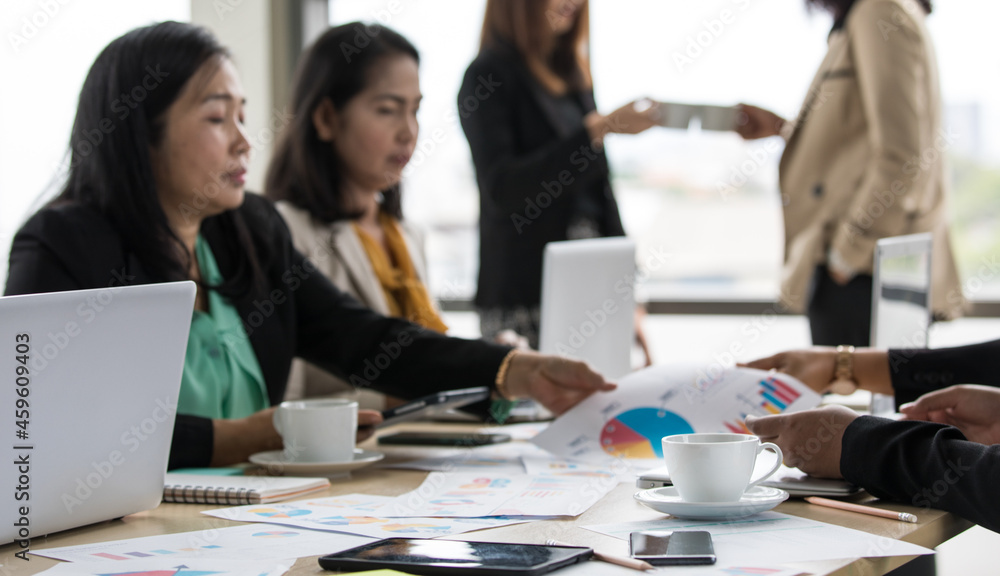 Fototapeta premium Close up shot of white hot coffee cup and dish on working meeting desk while unidentified unrecognizable businesswoman in casual business clothing working and cheers celebrating in blurred background