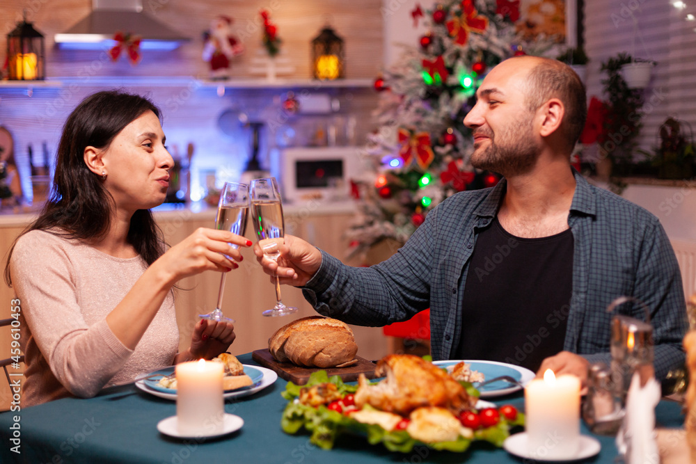 Happy family clinking glass of wine sitting at dining table celebrating christmas traditional holiday. Romantic cheerful couple spending christmastime together enjoying winter season Happy family clinking glass of wine sitting at dining table celebrating christmas traditional holiday. Romantic cheerful couple spending christmastime together enjoying winter season
