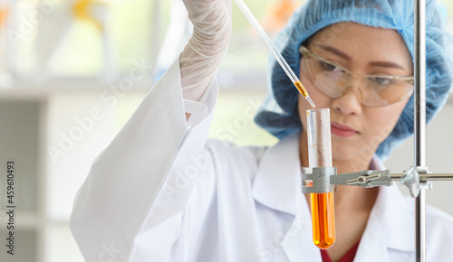 Asian female professional scientist in white lab coat safety goggles hygiene cap and rubber gloves holding looking red reagent solution sample in test tube in laboratory full of science equipment