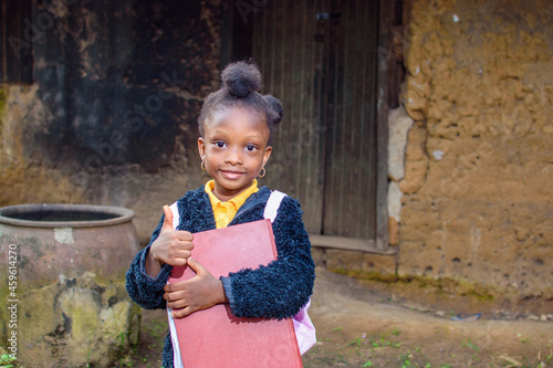 A happy little African girl child or student with pink school bag, holding and hugging her books outside a village mud house