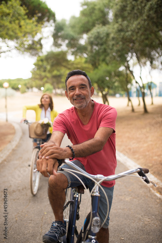 Portrait of happy mature riding bike in park, with his wife in background. Man resting in park, smiling at camera. Active seniors concept