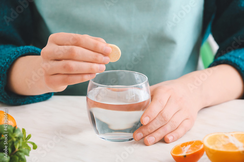 Woman putting ascorbic acid pill in glass of water on table in kitchen