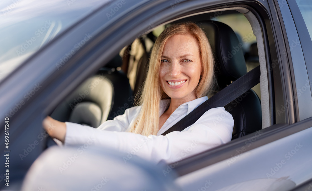 Portrait of smiling mature lady in formal wear sitting inside luxury car and keeping hands on steering wheel. View from lowered glass.