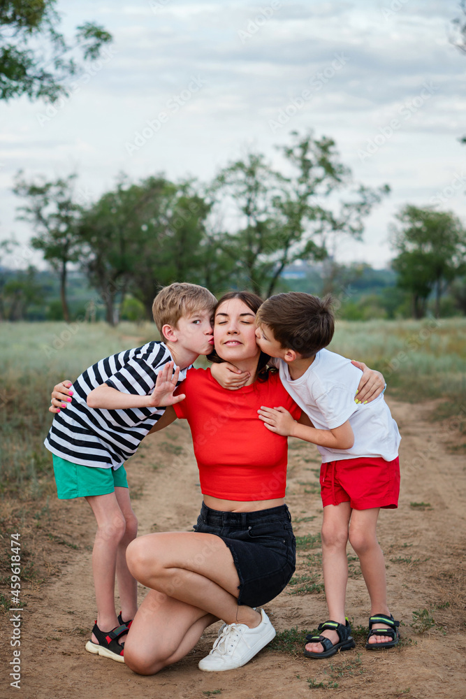Fototapeta premium Full-length authentic family portrait. Young casual mom with two sons hugging and kissing. Mother and child walking in countryside. Daily lives. Everyday activities.