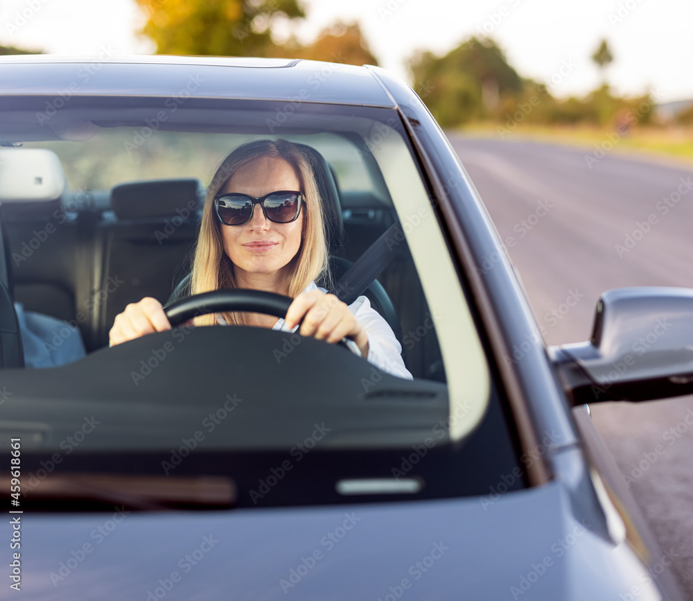 Confident mature lady with blond hair sitting inside modern car and keeping hands on steering wheel. Female driver wearing sunglasses and formal blue shirt.