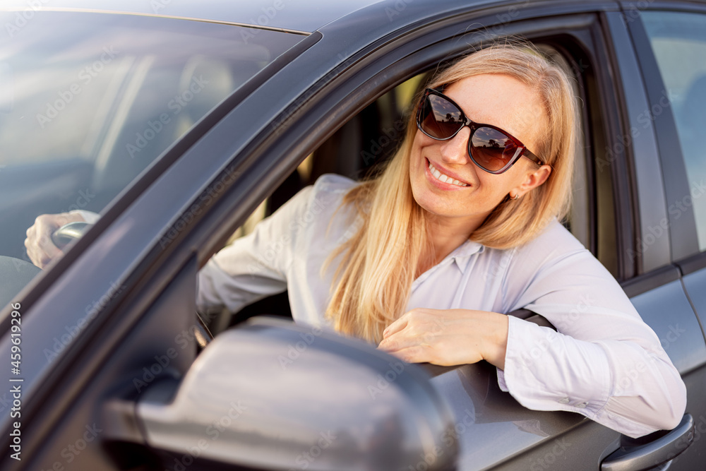 Smiling female driver in sunglasses sitting inside modern car and looking out of lowered glass. Concept of people, transport and positive emotions.