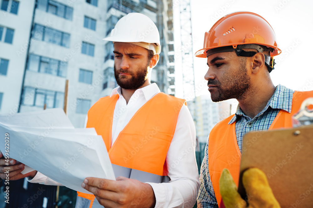 Two young male engineers in uniform and hardhats working at construction site