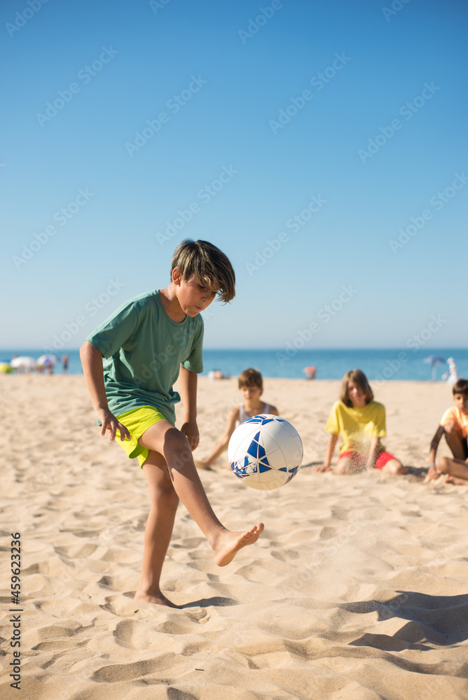 Concentrated preteen boy playing with ball on sand, his friends sitting and looking at him. Group of multiethnic friends on beach. Summer vacation concept
