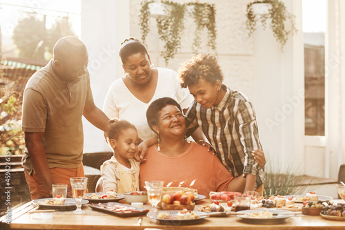 Portrait of happy African-American grandmother celebrating Birthday with family during dinner outdoors, lit by sunlight