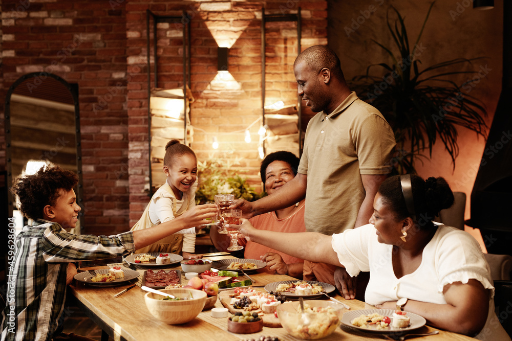 © Seventyfour - Warm toned portrait of happy African-American family clinking glasses and toasting while enjoying dinner together at evening