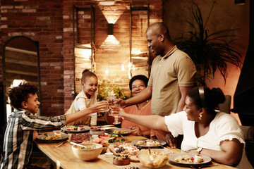 Warm toned portrait of happy African-American family clinking glasses and toasting while enjoying dinner together at evening