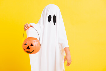 Funny Halloween Kid Concept, little cute child with white dressed costume halloween ghost scary he holding orange pumpkin ghost on hand, studio shot yellow on white background