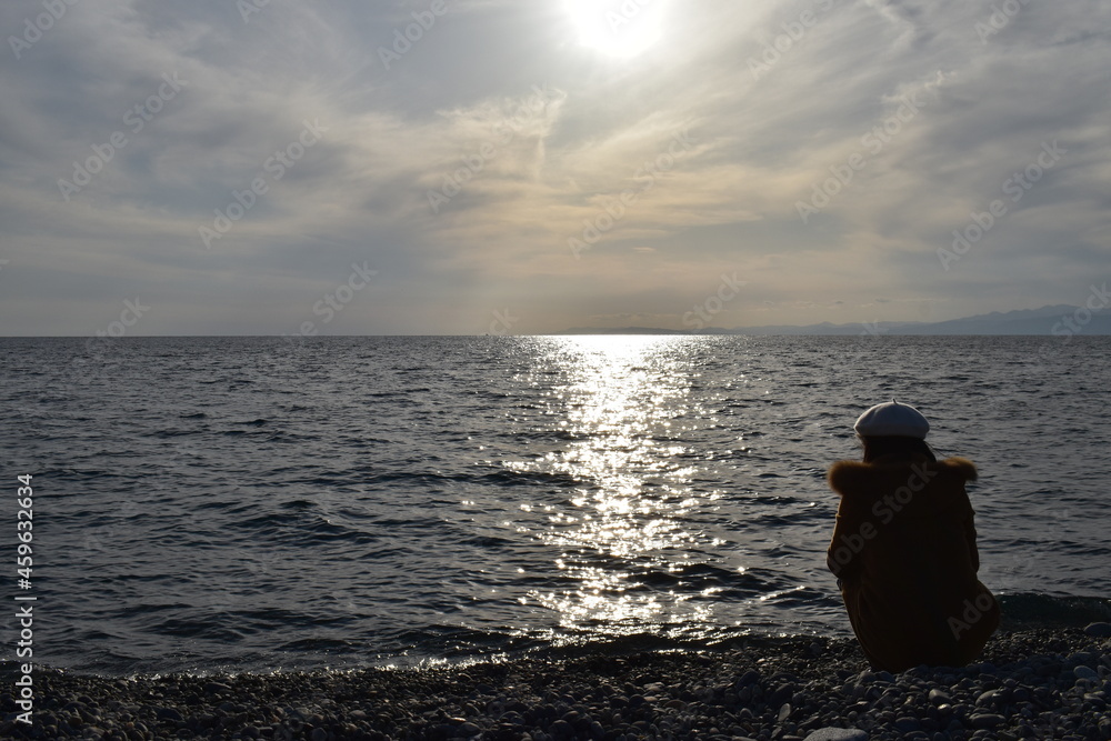 銀色の太陽と海と石の海岸にしゃがむ女性、白い帽子と茶色のコート