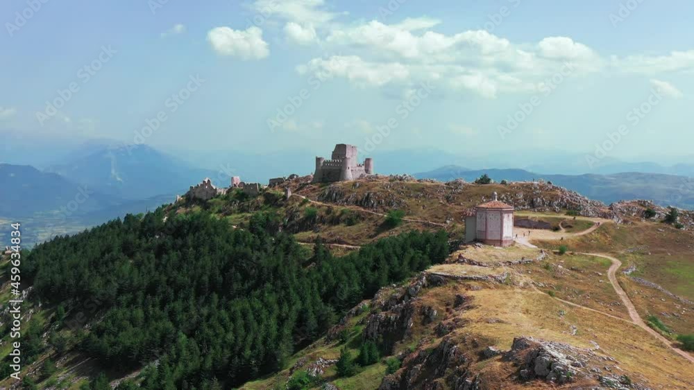 Aerial view of ancient castle on mountain hill, pine forest on mountain slope.White rocks and curved dirty road along the mountain. Mountain range in the background with blue sky un a summer sunny day Aerial view of ancient castle on mountain hill, pine forest on mountain slope.White rocks and curved dirty road along the mountain. Mountain range in the background with blue sky un a summer sunny day
