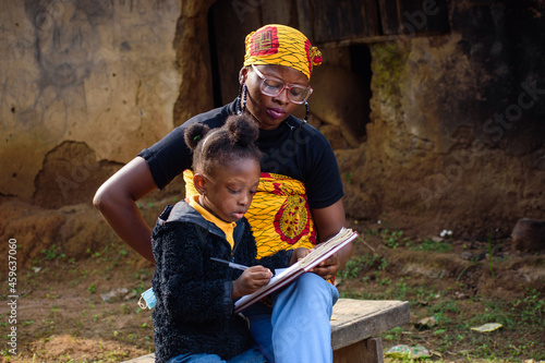 An African mother wearing glasses with a baby on her back, sits outside a village mud house, helping a girl wearing nose mask with studies for excellence in her school, career and education